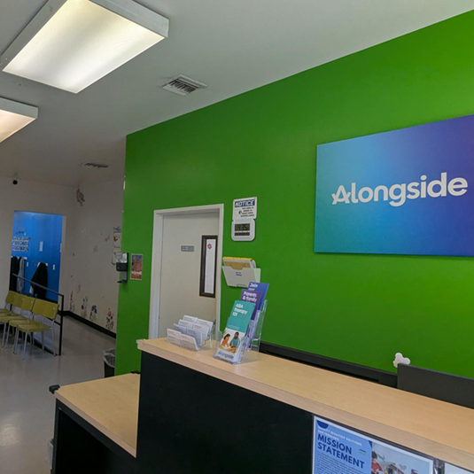 The San Fernando clinic's front desk, with a bright green accent wall, a blue Alongside sign, a brochure rack, and a waiting area with yellow chairs.