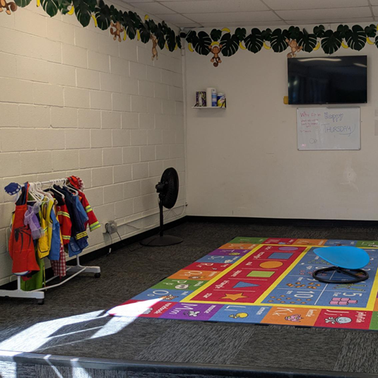 Group learning room with a dress-up costume rack, a colorful numbers-and-shapes floor mat, a wall-mounted screen, a whiteboard, and a jungle-vine border.