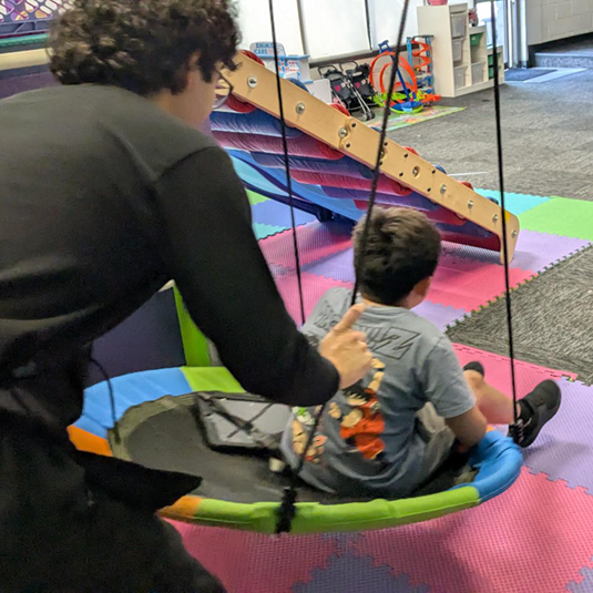 A therapy session on a platform swing in the sensory gym, with interlocking foam mats and a wooden climbing ramp in the background.