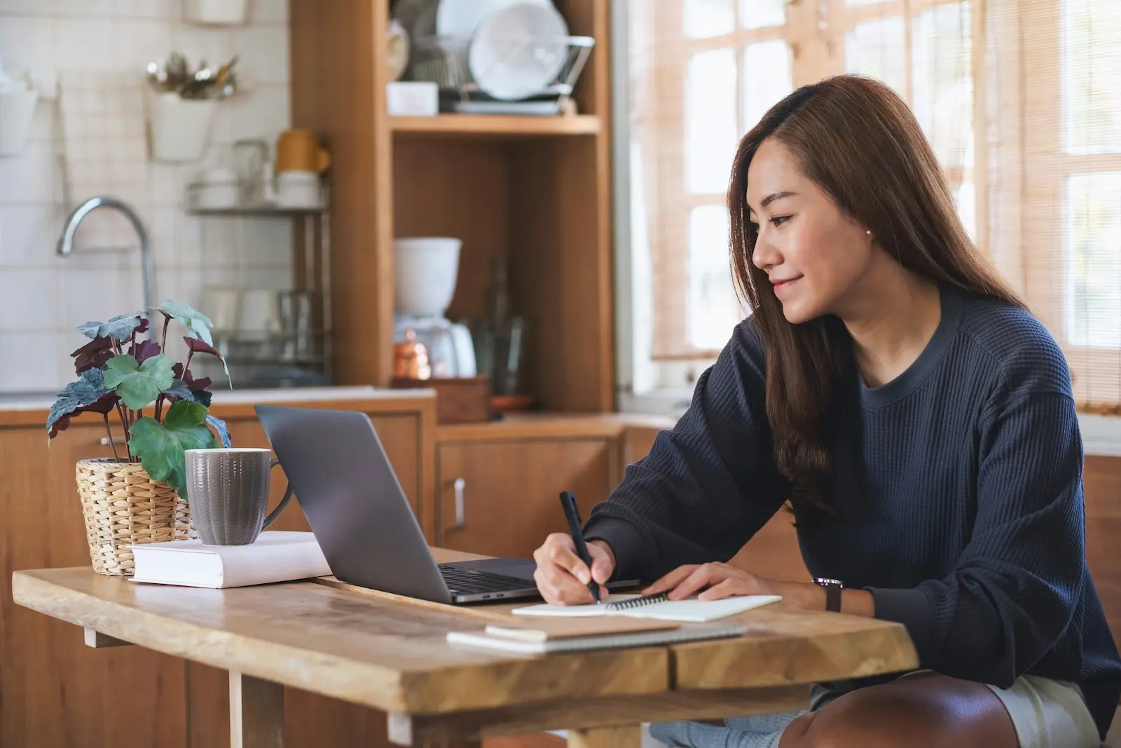 Real estate investor taking notes next to her laptop while managing her LLC paperwork from her kitchen table