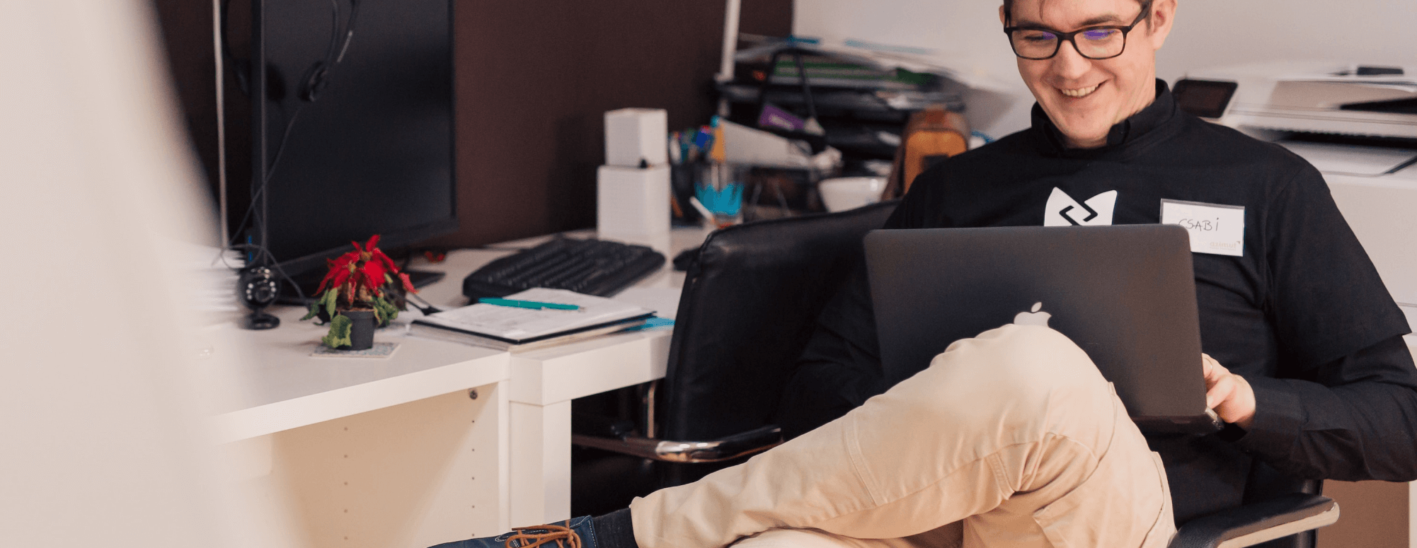 Smiling man wearing glasses working on a laptop in an office with a desk and computer in the background.