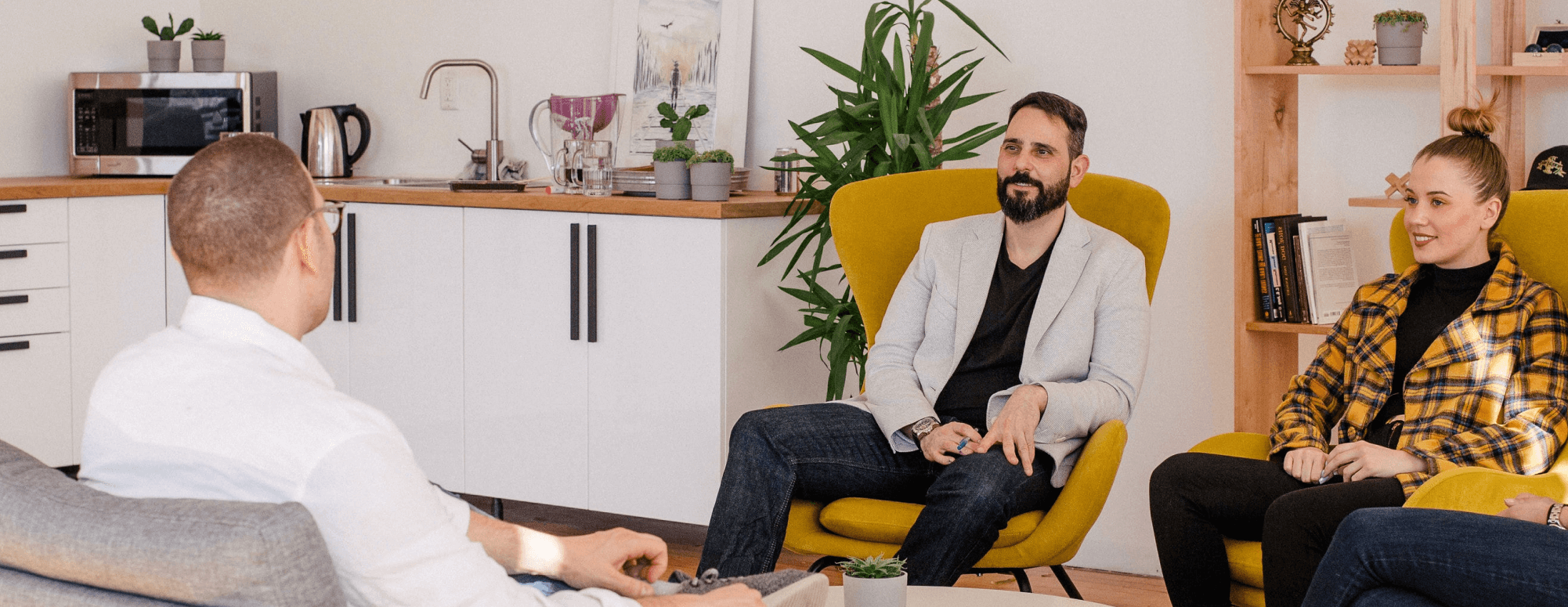 Three people seated in a casual office setting having a discussion, with kitchen and plants in background.