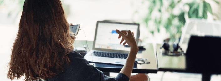 Person with long hair using a smartphone and laptop at a glass table indoors with plants in the background.