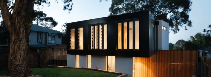 Modern two-story house with large vertical windows glowing at dusk in a wooded neighborhood.