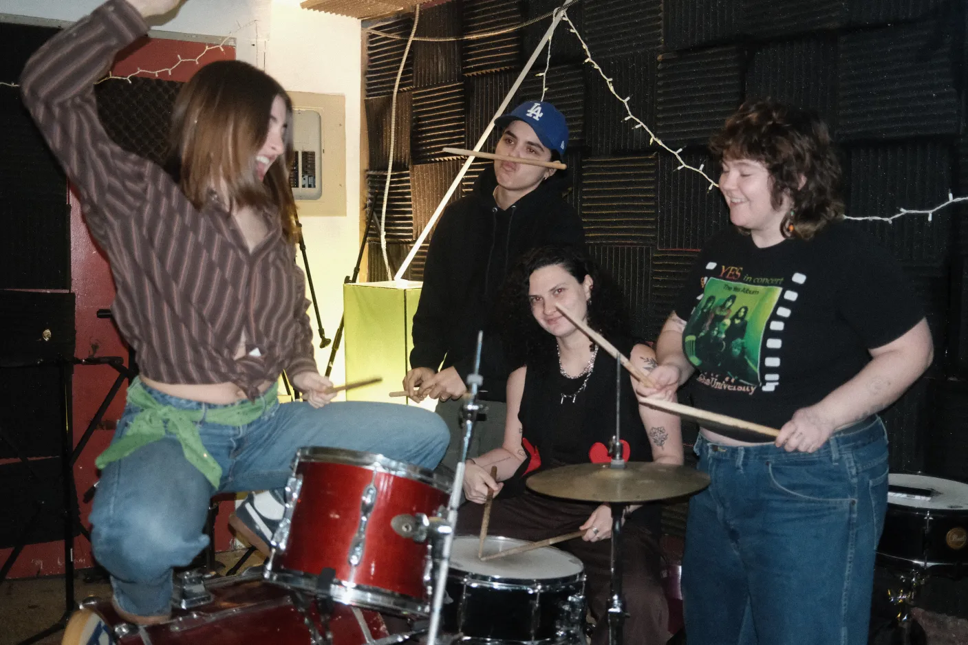 The four members of talon gathered around a drum set in a basement rehearsal space. They're being silly and goofy and twirling sticks