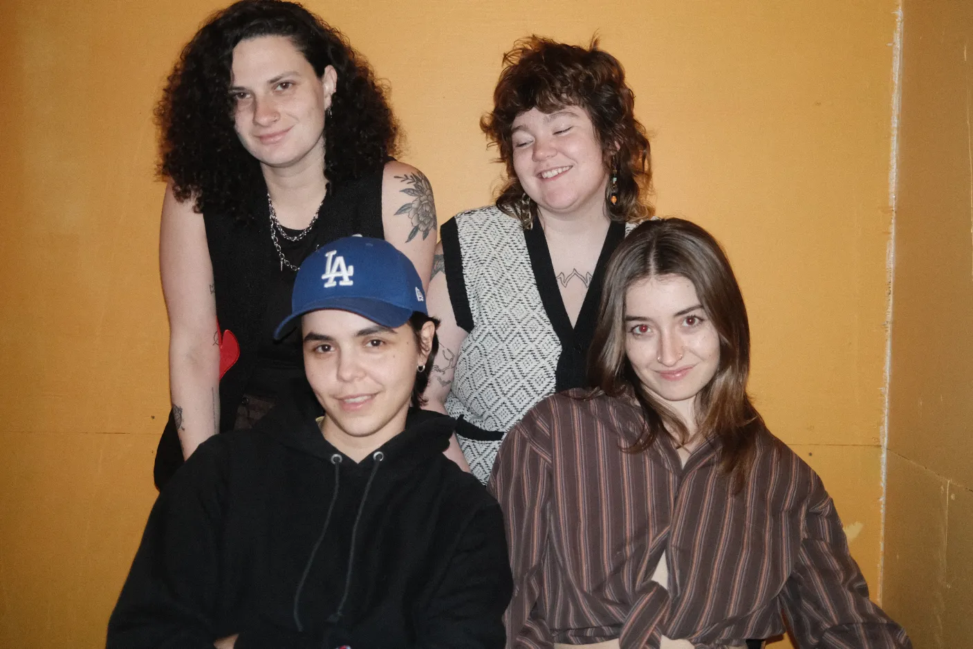 The four members of Talon against an orange wall. They're all kind of smirking or laughing in their own way. 