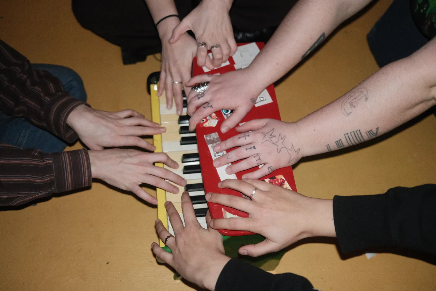 Four pairs of hands, one with tattoos, all belonging to the four members of the band Talon, reaching towards a children's keyboard