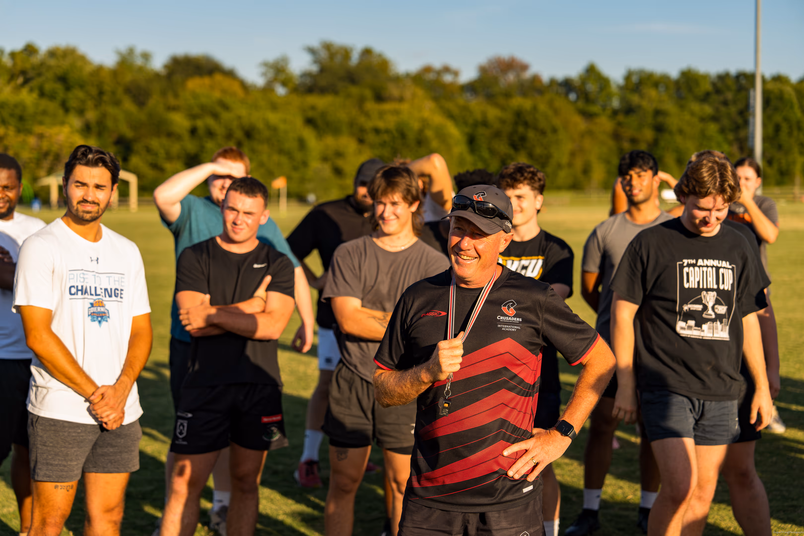 Coach wearing a black and red Crusaders shirt and cap smiles while standing with a group of young athletes on a sunny sports field.