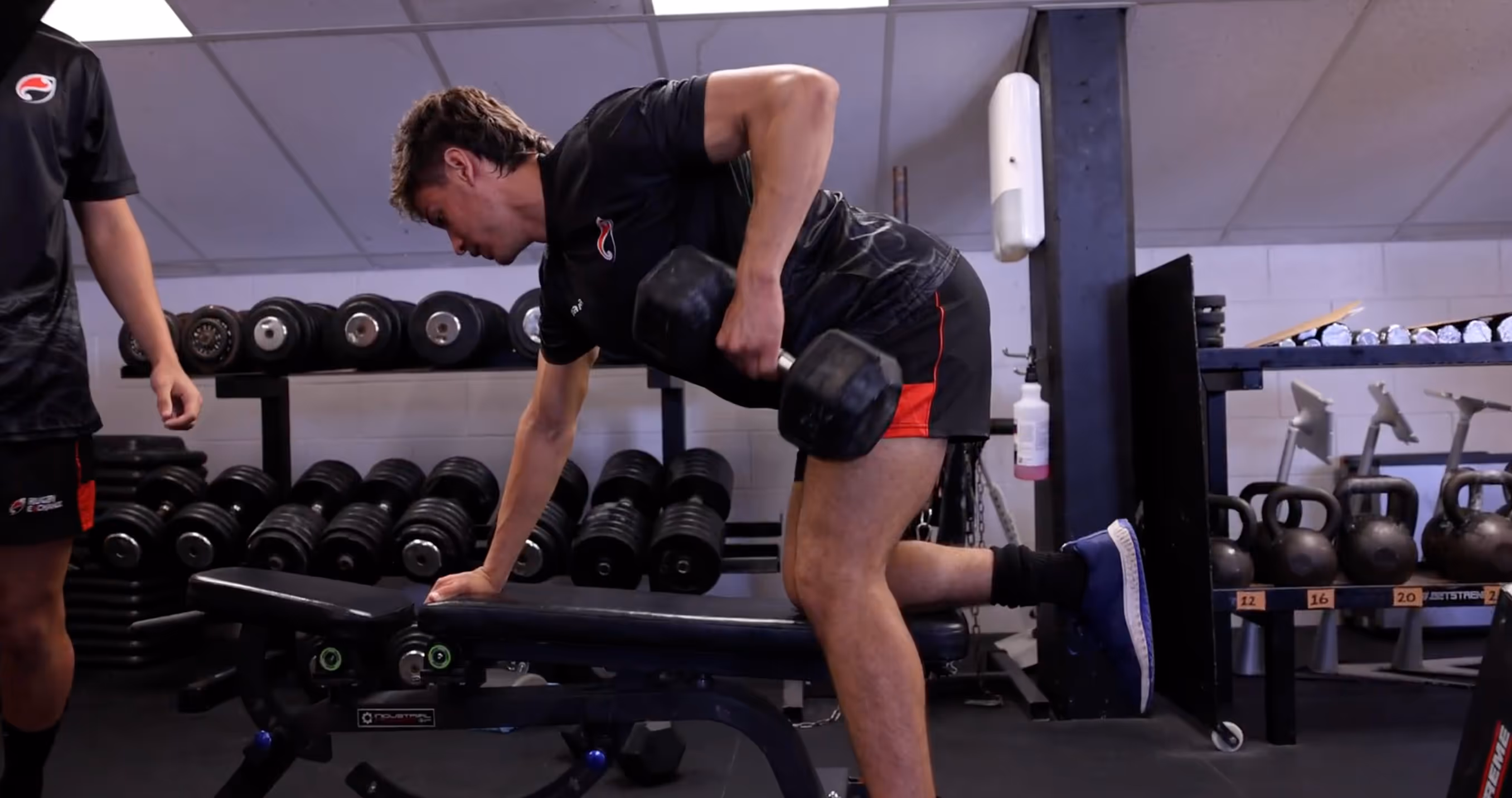 Man performing single-arm dumbbell row exercise with one knee on a bench in a gym.