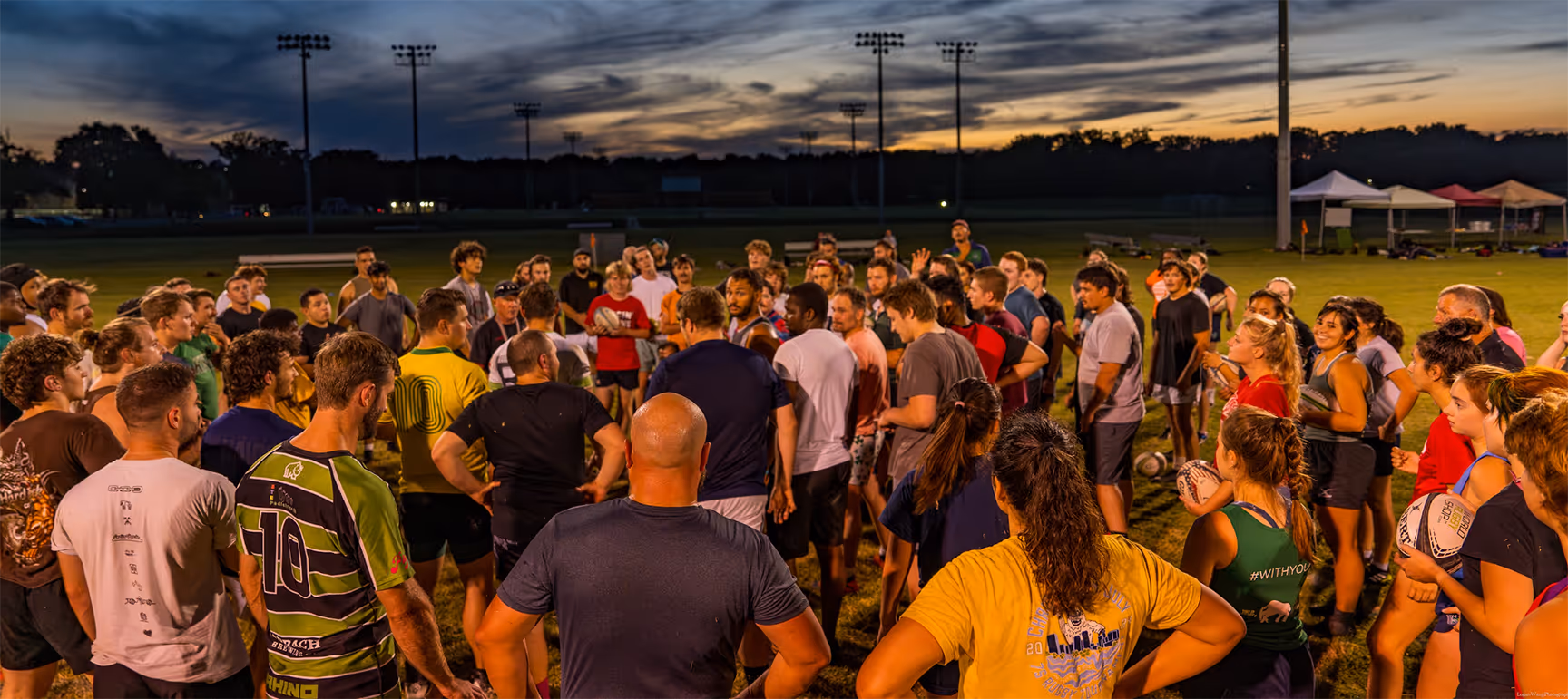 Large group of people gathered on a sports field at dusk, some holding rugby balls, listening to a person speaking in the center.