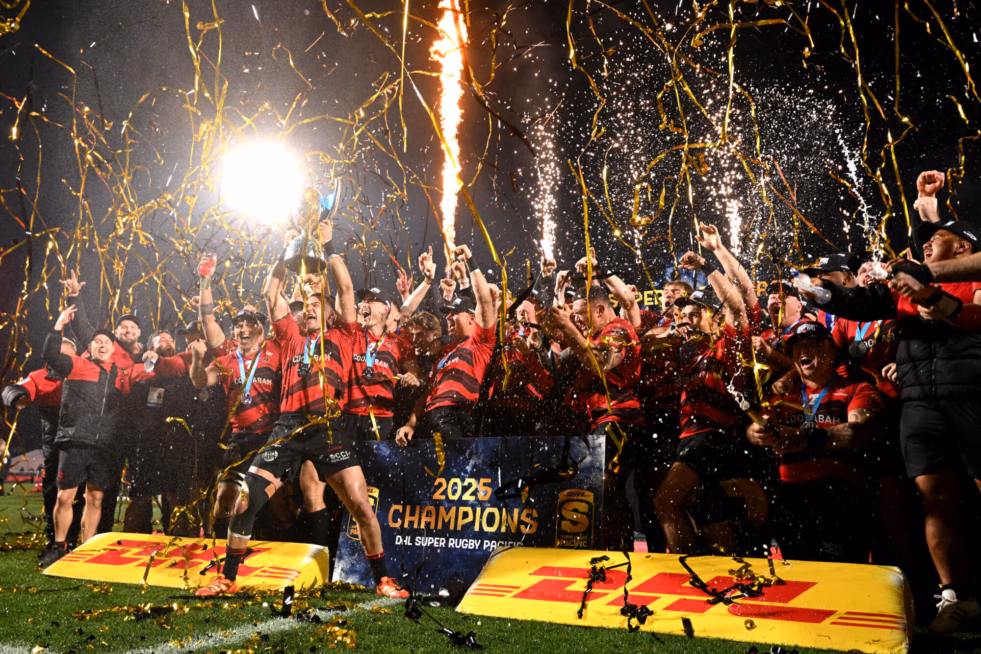 Rugby team in red and black uniforms celebrating with a trophy, confetti, and fireworks on a field with a '2025 Champions DHL Super Rugby Pacific' sign.