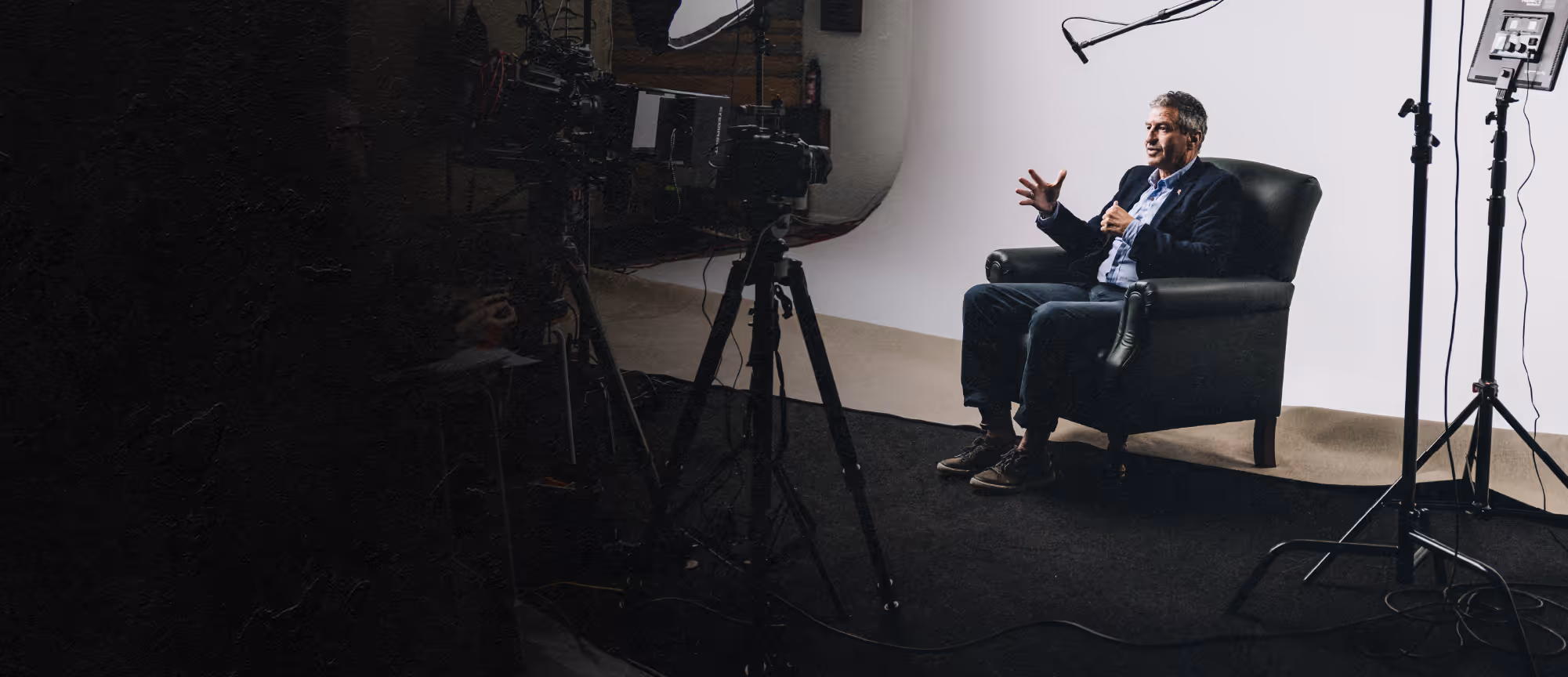 Man in a dark suit sitting in a black armchair, gesturing during a video interview in a studio with cameras and lighting equipment.