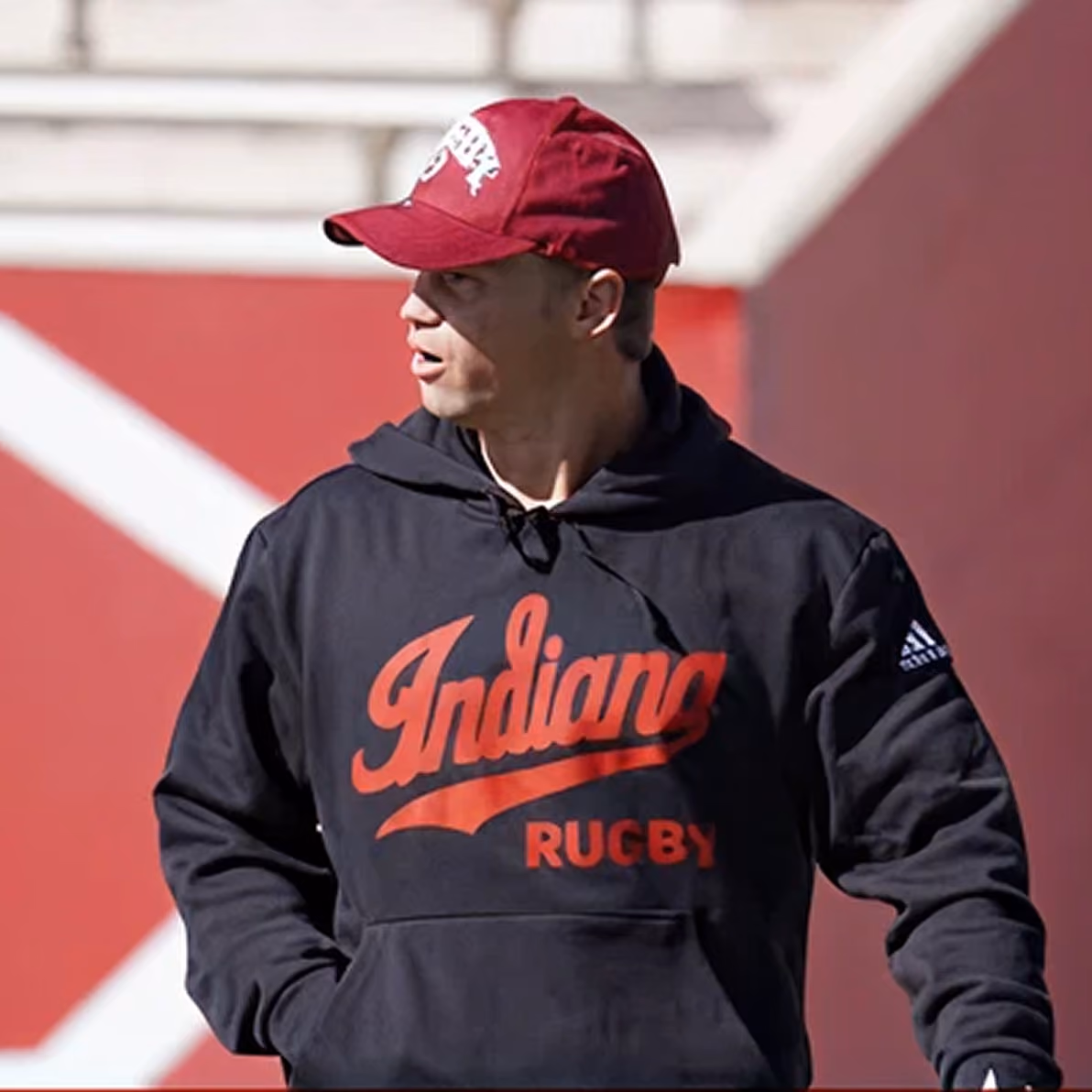 Man wearing an Indiana Rugby hoodie and a red cap looking to the side outdoors.