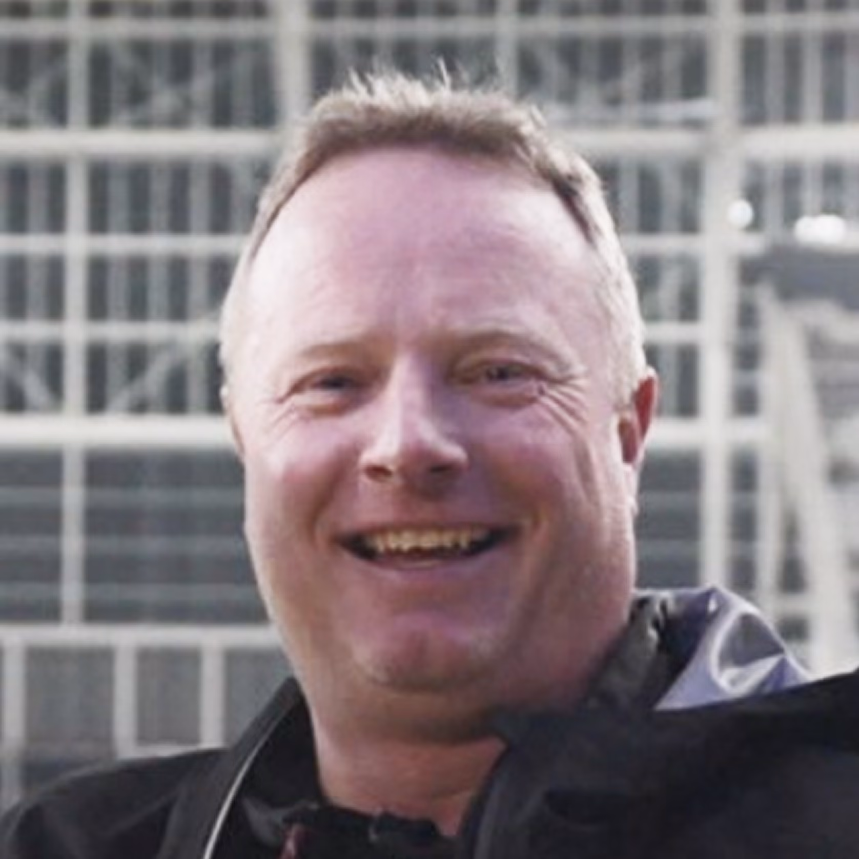 Smiling middle-aged man with short light hair wearing a dark jacket, outdoor background.