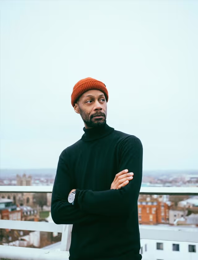 A man wearing a black sweater and an orange beanie stands on a balcony, looking out at the view.