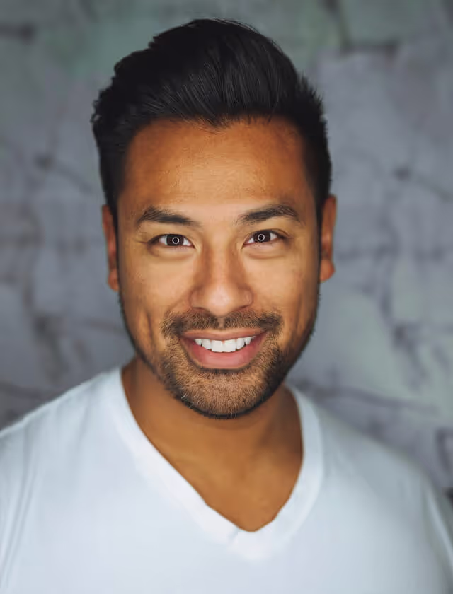 A man wearing a white shirt with black hair, standing confidently against a neutral background.