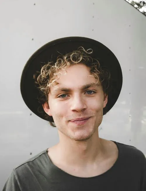 A young man with curly hair is wearing a stylish hat, exuding a casual and confident demeanor.