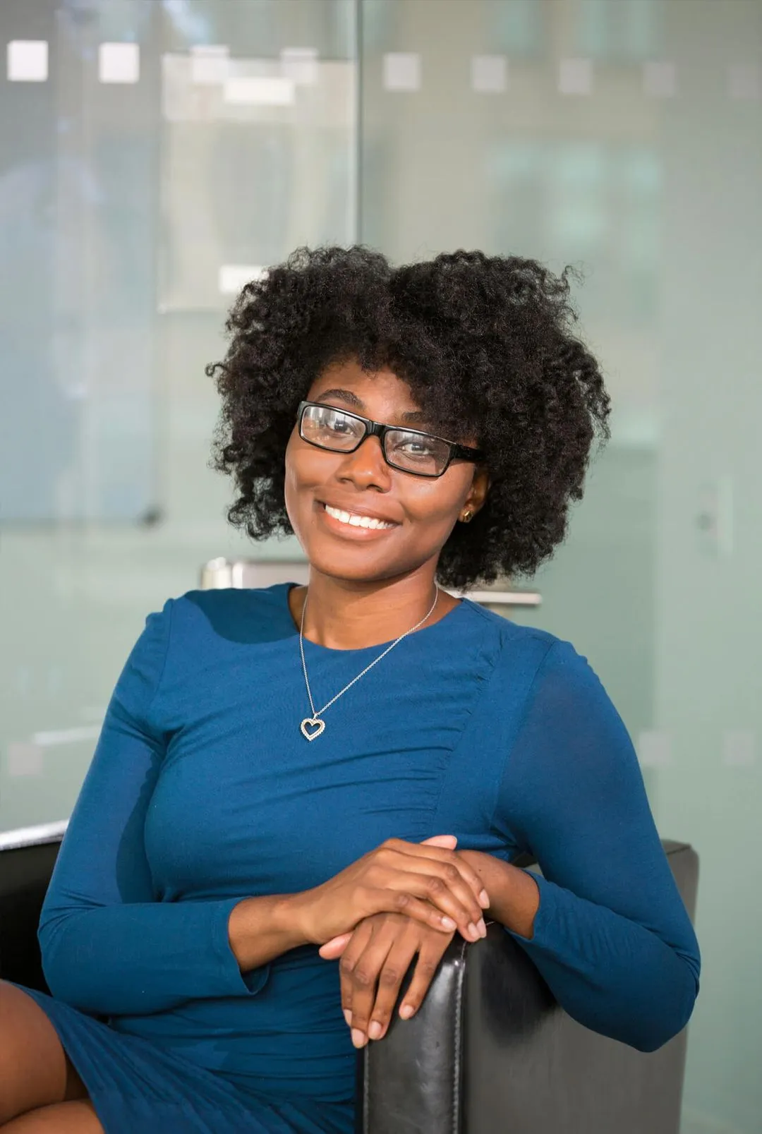 A woman wearing glasses and a blue dress is seated on a chair, exuding a calm and composed demeanor.