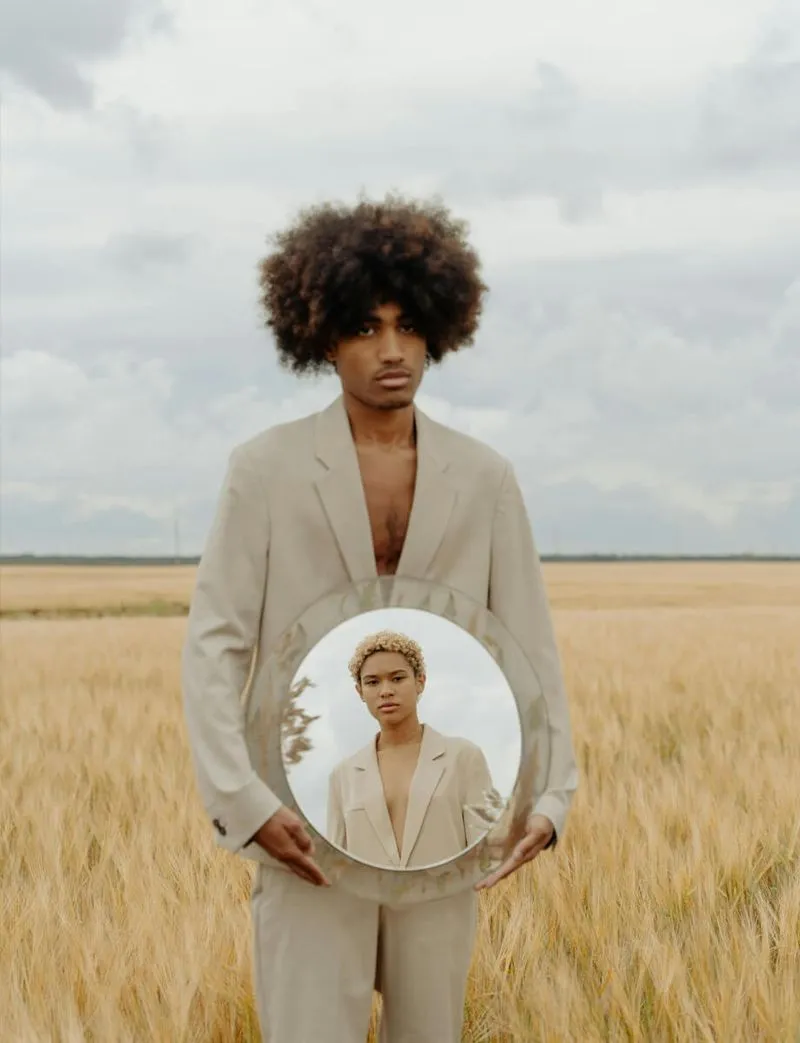 A man in a suit stands in a field, holding a mirror that reflects the surrounding landscape.