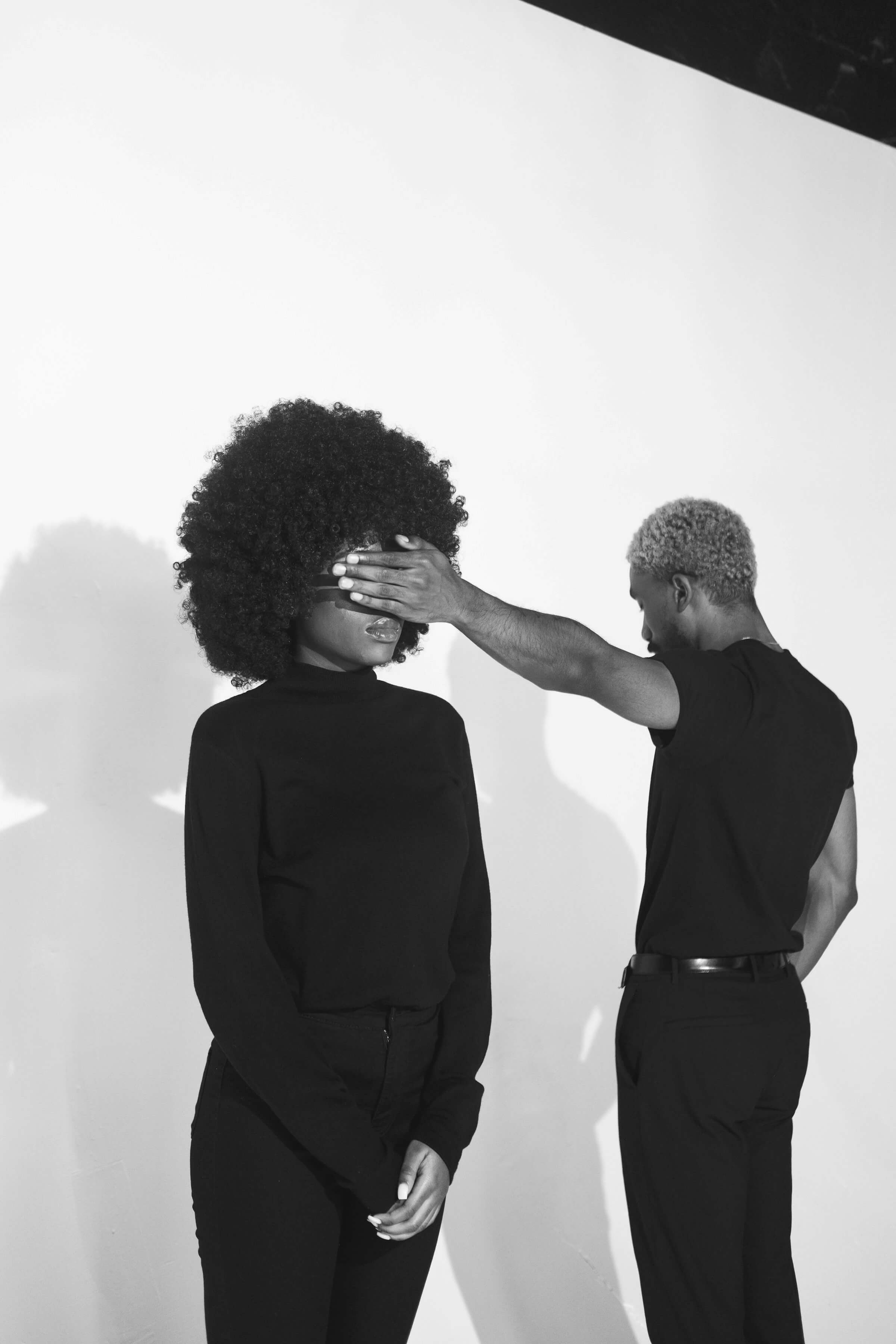 A man and woman stand together in front of a plain white wall, both smiling and dressed casually.