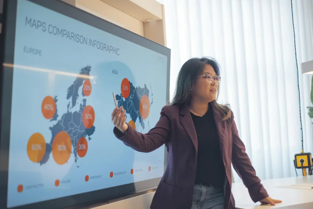 A woman in a blazer presents a maps comparison infographic on a screen, highlighting data for Europe and Asia with percentages in circles.