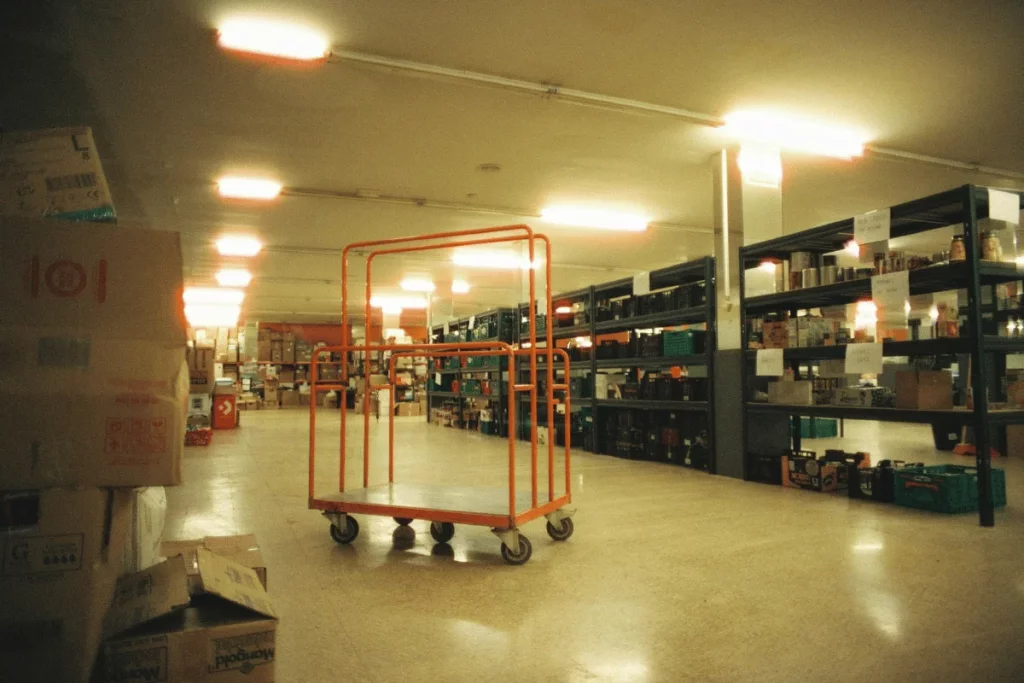 Warehouse aisle featuring inventory boxes, shelving racks, and a central orange dolly.