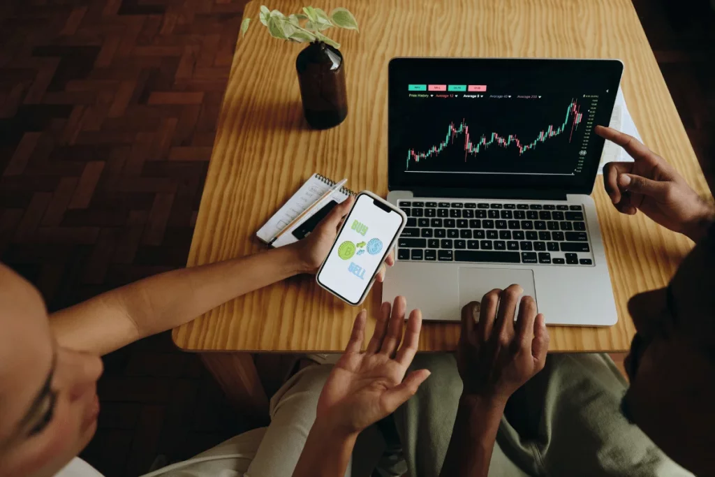 Two individuals analyzing stock market trends on a laptop with a smartphone displaying buy and sell options.