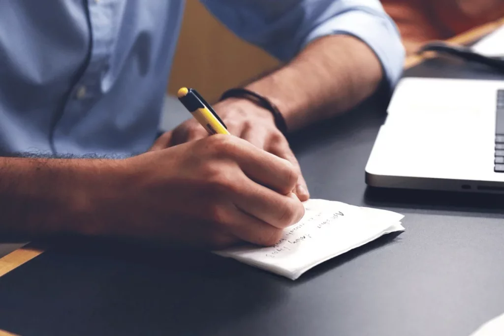 Close-up of a person jotting down notes on a post-it beside a laptop.
