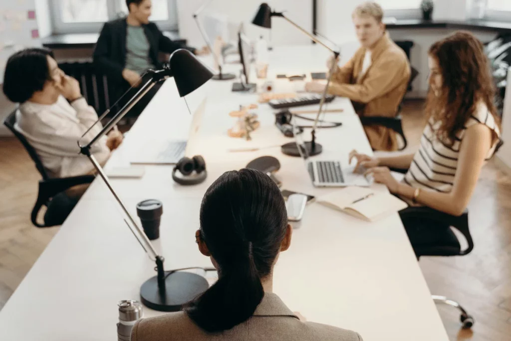 A group of five people collaborating in a modern office space, working at desks with laptops and various office equipment.