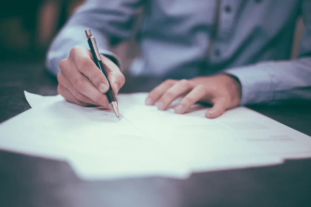 Detailed view of a person's hand signing a paper with a pen on a desk with a dark, blurred background.