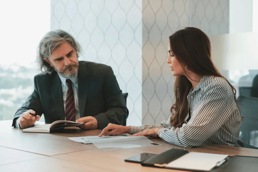Professional meeting between a young woman and an older man reviewing paperwork at a desk.