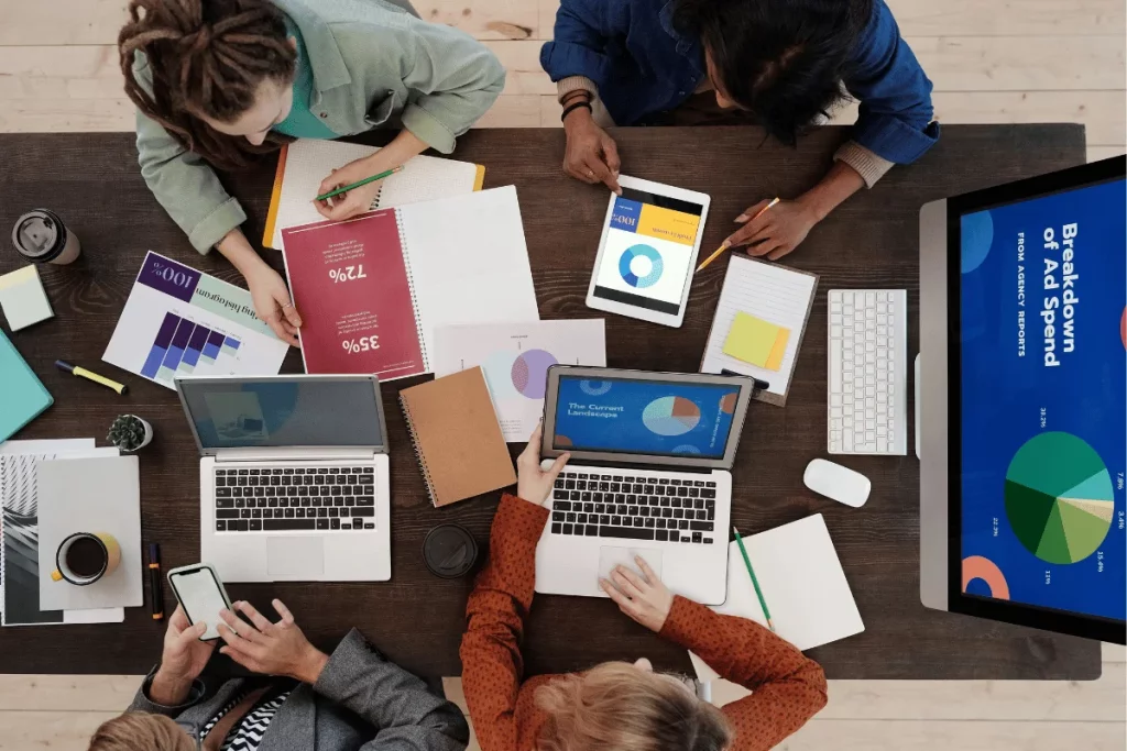 Team collaborating on marketing strategy with laptops, tablets, and printed reports.