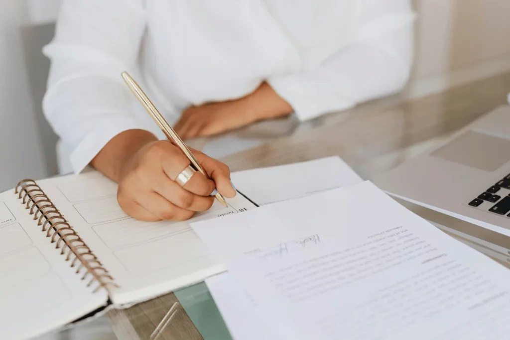 Person writing in a planner with documents and a laptop on the desk.