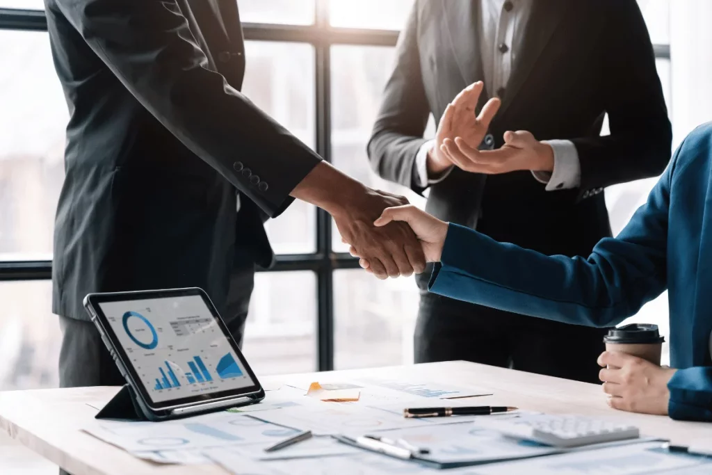 Business professionals shaking hands during a meeting with data charts on a tablet in the foreground.