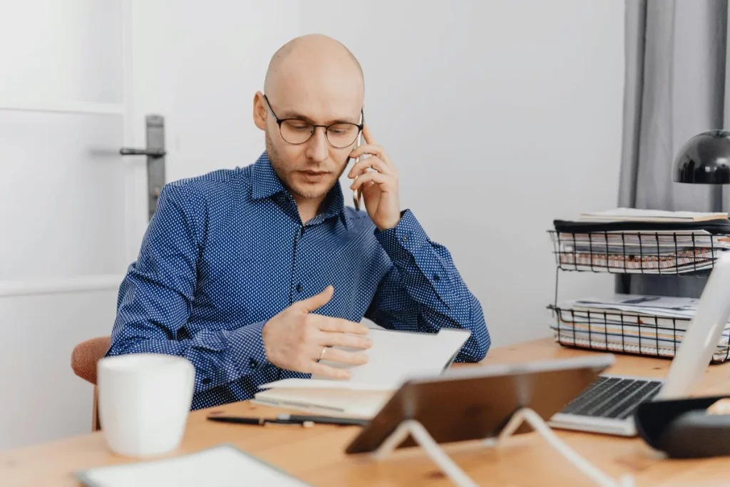 A bald man in glasses talking on his phone while looking at documents at his desk.