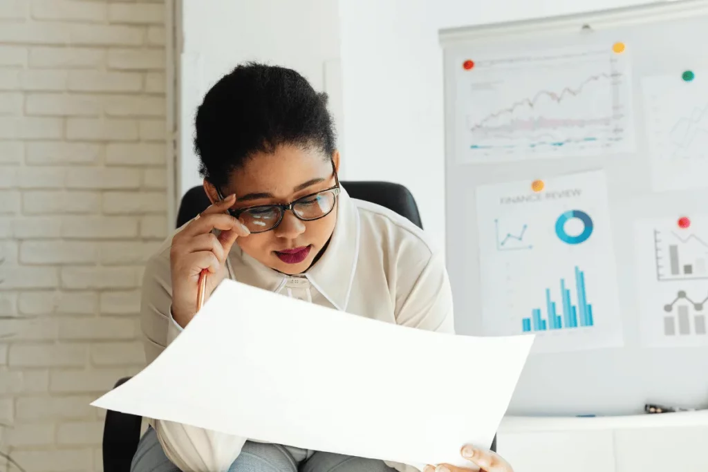 Businesswoman reviewing financial documents in an office with charts and graphs displayed on a whiteboard in the background.