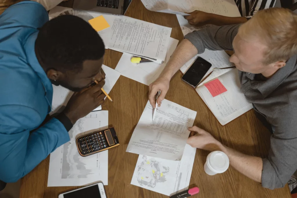 Two colleagues reviewing documents at a table filled with notes, a calculator, and a coffee cup during a business meeting.