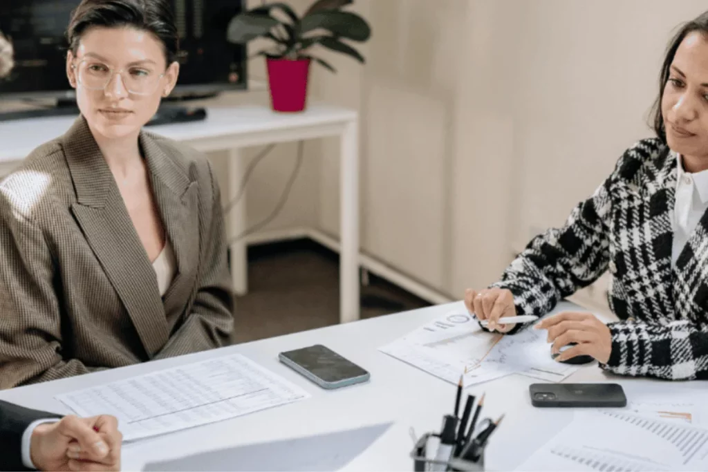 Two businesswomen in a meeting, reviewing documents and charts at a white table in a modern office.