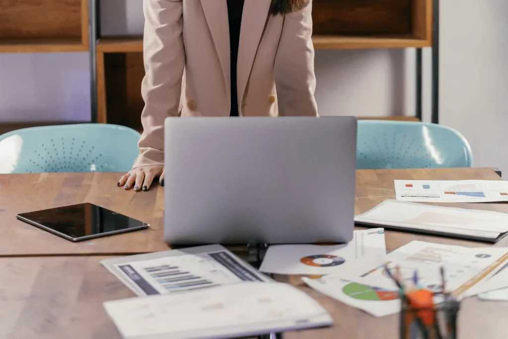 Businesswoman in a beige blazer leaning on a wooden table with a laptop and tablet.
