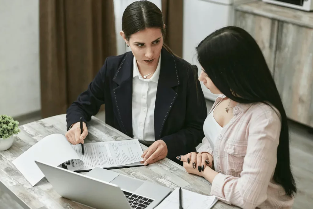 Two women discussing paperwork at a table with a laptop.