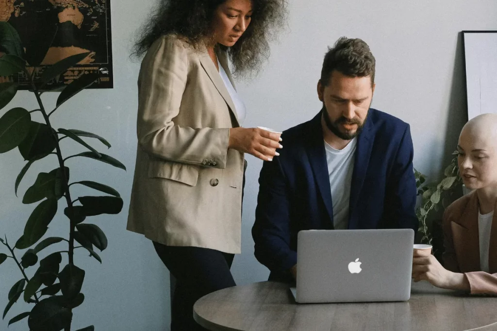 Office meeting with three people, one using a laptop, and two others standing and discussing.
