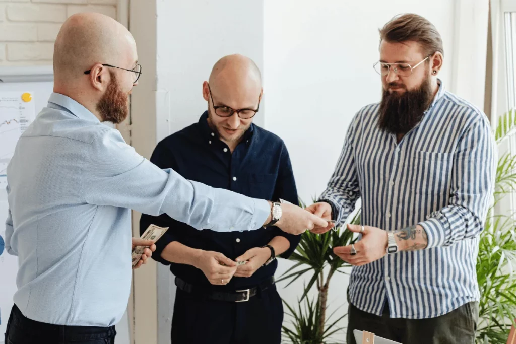 Three men exchanging dollar bills in a business meeting, standing near a whiteboard with charts.