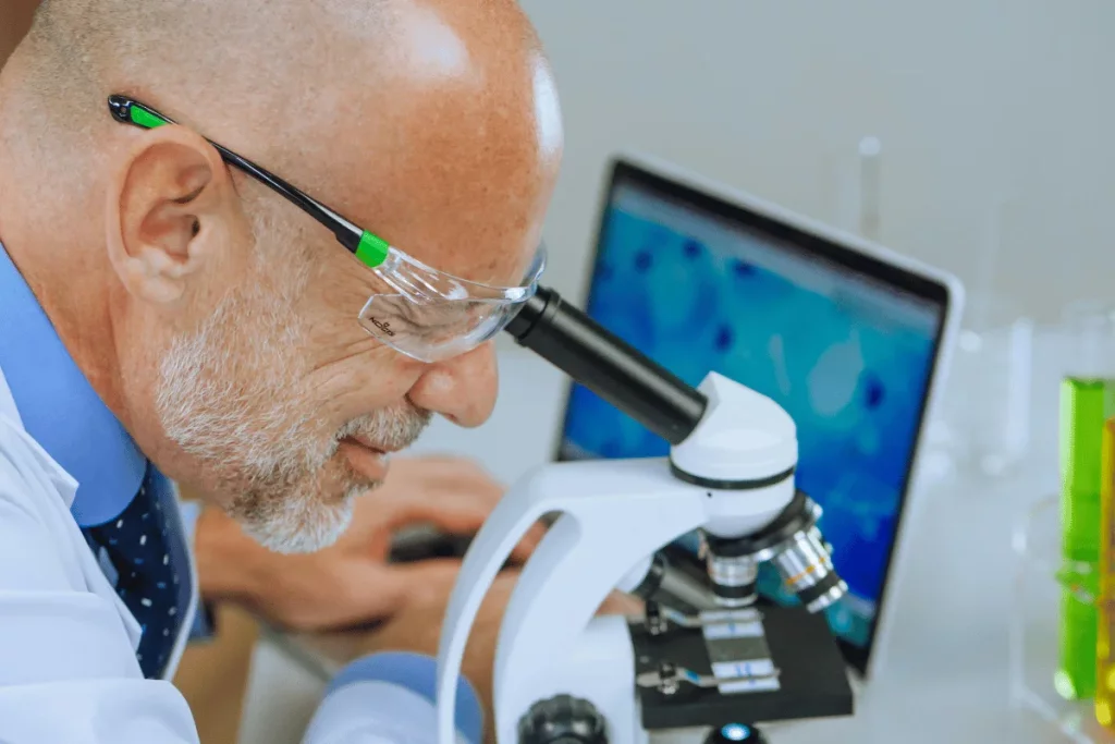 A scientist in a lab coat examines samples under a microscope, with colorful solutions in beakers and a computer displaying data nearby.
