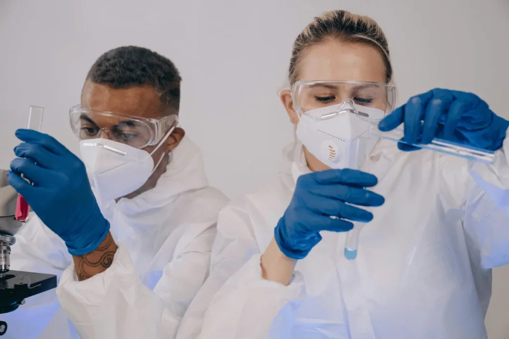 Two scientists in protective gear conduct experiments, one holding a test tube with red liquid and the other with a pipette.