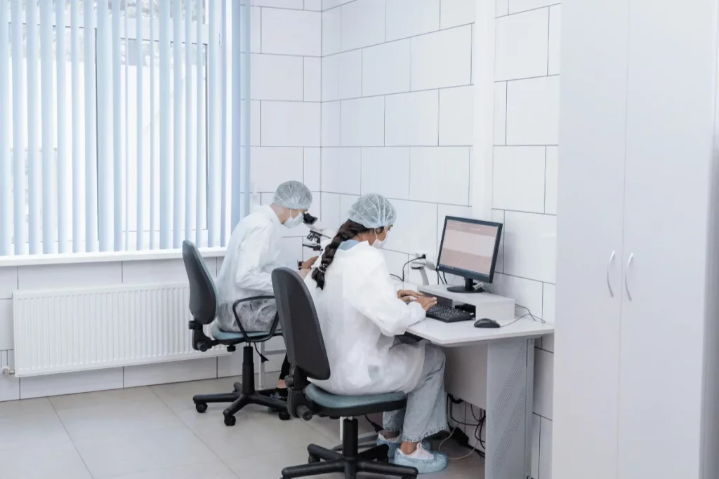 Two lab technicians working in a sterile laboratory using a microscope and computer.