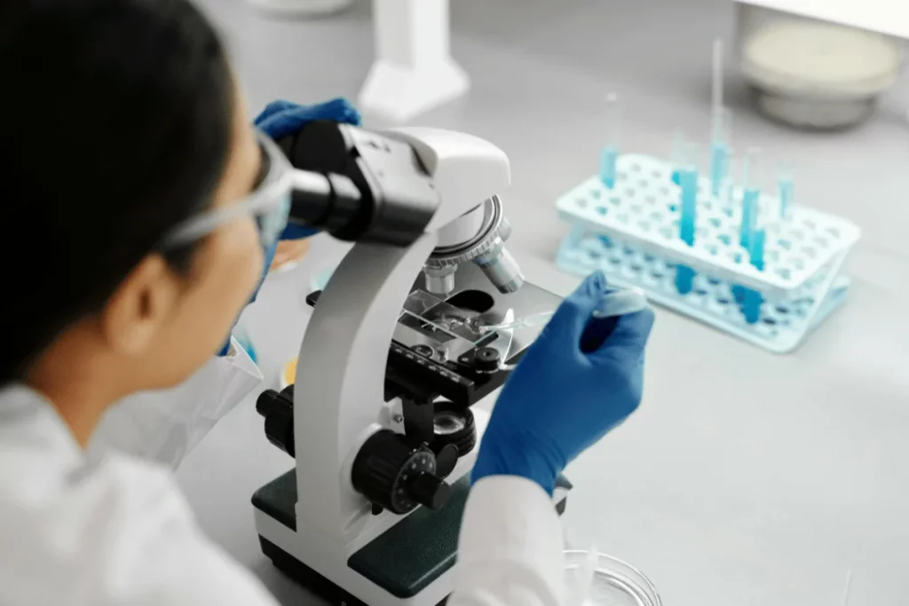 A person in a lab coat and blue gloves uses a microscope, preparing a slide with a pipette, surrounded by lab equipment.