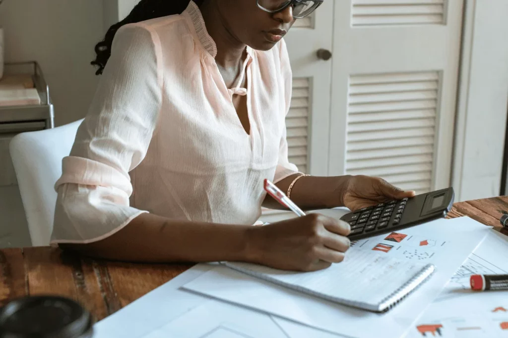A woman in a pink blouse uses a calculator while taking notes and analyzing documents on a wooden desk.