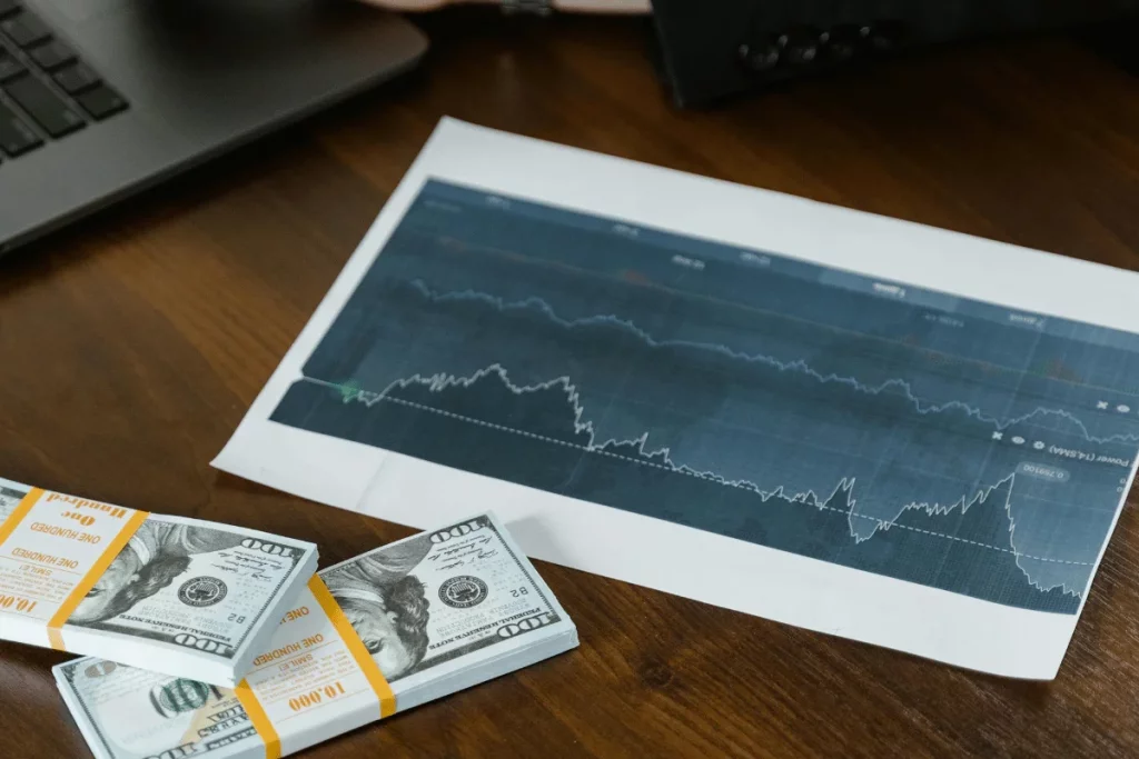 A stack of $100 bills beside a printed stock market graph on a wooden table, with a laptop partially visible in the background.