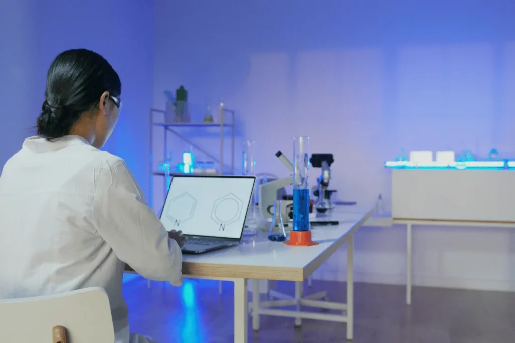 A scientist in a lab coat analyzes data on a laptop, surrounded by beakers and scientific equipment in a blue-lit laboratory.
