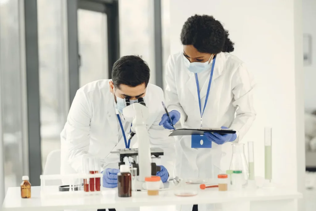 Two scientists in lab coats examine samples under a microscope amid various glassware and containers filled with liquids on a table.
